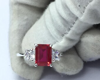 Peut inclure: Bague en argent ornée d'une grande pierre rouge rectangulaire, flanquée de deux petites pierres claires carrées. L'anneau est présenté sur fond blanc.