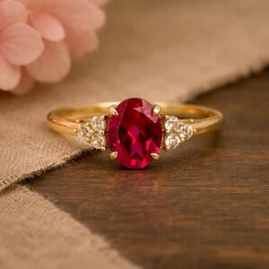 May include: A gold ring featuring a large, oval-cut, deep red gemstone at the centre, held by four prongs. On each side of the central stone, there is a cluster of three small, clear, round-cut accent stones set into the polished gold band.