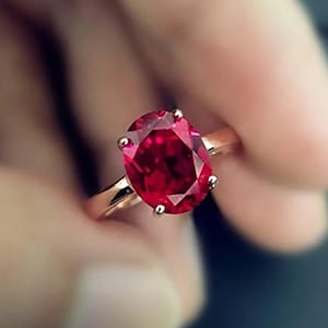 May include: A gold ring featuring a large, oval-cut, deep red gemstone. The gemstone is held in place by four prongs, and the band is a simple, polished gold. The ring is shown in a close-up view.