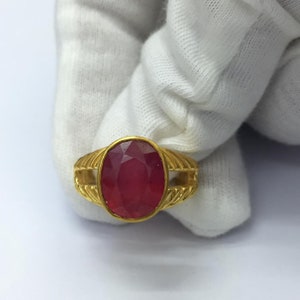 Op de afbeelding: Een gouden ring met een ovale, dieprode edelsteen. De ring heeft een getextureerd, geribbeld ontwerp op de band en rond de edelsteen setting. De edelsteen is het middelpunt en toont zijn gefacetteerde oppervlak en rijke kleur.
