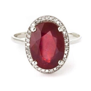 May include: A silver ring featuring a large, faceted, oval-shaped ruby gemstone. The ruby is surrounded by a halo of small, sparkling white gemstones. The ring's band is a simple, polished silver.