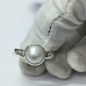 May include: A silver ring with a large, round, lustrous pearl. The pearl is set in a bezel setting, and the band is smooth and polished. The ring is held by a gloved hand against a white background.