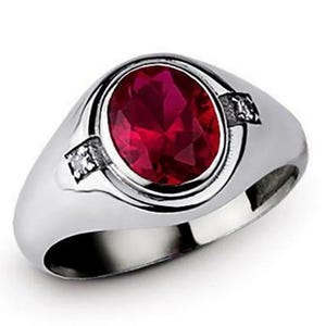 May include: A silver ring featuring a large, oval-cut, deep red gemstone as the centerpiece. Two small, clear accent stones are set on either side of the main stone. The ring has a polished, reflective surface.