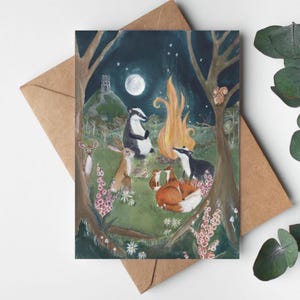May include: A greeting card featuring a whimsical illustration of woodland animals gathered around a campfire under a full moon. The scene includes a badger, fox, rabbit, and other creatures in a forest setting, with a castle in the background.