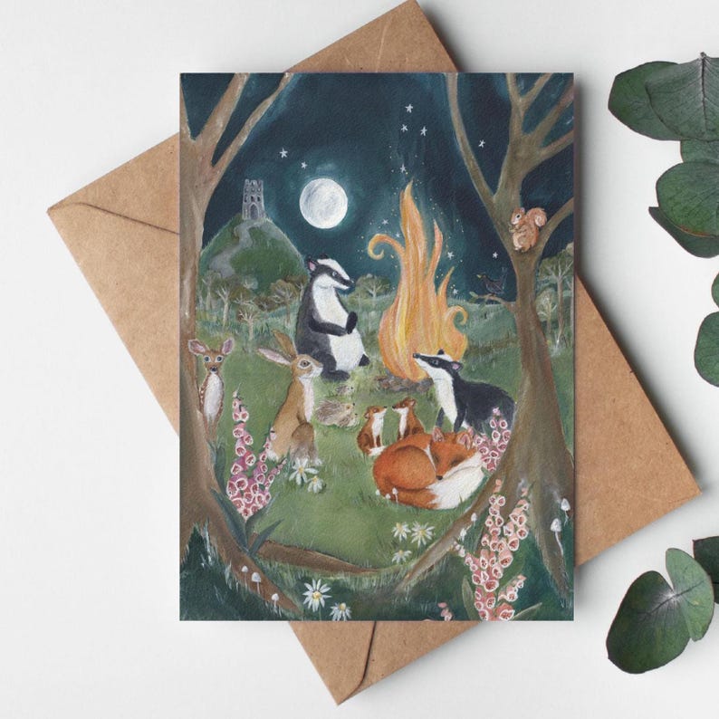 May include: A greeting card featuring a whimsical illustration of woodland animals gathered around a campfire under a full moon. The scene includes a badger, fox, rabbit, and other creatures in a forest setting, with a castle in the background.