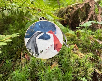 May include: A round wooden ornament featuring a badger and a fox nose to nose in a snowy scene under a starry night sky. The ornament has a black and white ribbon for hanging and is surrounded by green foliage.