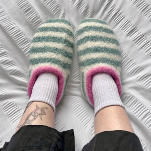 Puede incluir: Un par de zapatillas de lana a rayas en tonos turquesa y crema, con ribete rosa. Las zapatillas se llevan con calcetines grises. El tema general son calzado c&oacute;modo y acogedor.