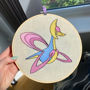 May include: Embroidered hoop art featuring a pink, blue and yellow mythical creature with a crescent moon on its head.