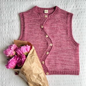 May include: A sleeveless, knitted vest in a dusty rose hue, featuring a round neckline and a series of wooden buttons down the front. A bouquet of pink peonies wrapped in brown paper sits beside the vest, creating a soft, feminine aesthetic. The vest is likely handmade.