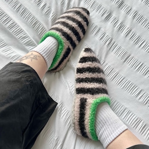 Puede incluir: Un par de zapatillas a rayas negras y beige, con un ribete verde brillante. Las zapatillas se llevan con calcetines blancos. La imagen muestra una vista de cerca de los pies y las piernas.