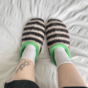 Puede incluir: Un par de zapatillas a rayas con rayas negras y beige, ribete verde y calcetines blancos. Las zapatillas est&aacute;n sobre una superficie blanca texturizada. La imagen muestra las piernas y los pies.