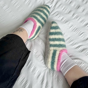Puede incluir: Un par de zapatillas de lana a rayas en tonos turquesa, blanco y rosa. Las zapatillas se llevan con calcetines blancos y grises. Las zapatillas est&aacute;n sobre un edred&oacute;n blanco texturizado. La persona lleva pantalones negros.
