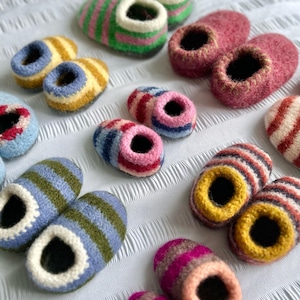 以下が含まれることがあります： カラフルな手作りのウールスリッパのコレクション。スリッパは、ピンク、緑、青、黄色、赤の色合いで、さまざまなストライプ模様が特徴です。各スリッパは、開口部の周りにコントラストの白いトリムが付いています。