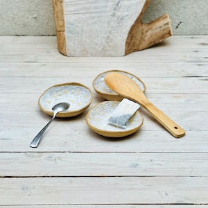 Könnte beinhalten: Drei kleine weiße Keramikschalen mit gelbem Gänseblümchenmuster, ein Holzlöffel und ein Teebeutel. Die Schalen stehen auf einer weißen Holzoberfläche.