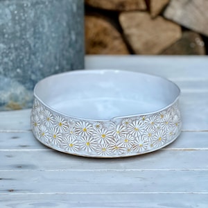 May include: A white ceramic bowl with a raised daisy pattern around the rim. The daisies have yellow centers.