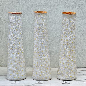 Op de afbeelding: Drie witte keramische vazen met een geel madeliefjespatroon. De vazen zijn hoog en slank met een uitlopende bovenkant. De vazen zijn genummerd 1, 2 en 3.