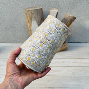 Op de afbeelding: Een witte keramische vaas met een geel madeliefjespatroon. De vaas is handgemaakt en heeft een getextureerd oppervlak.
