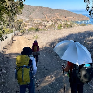 May include: Four people hike on a dirt path with backpacks and hiking poles. The path leads to a small town in the distance. One person is holding a silver umbrella.
