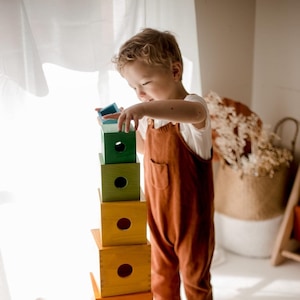 May include: A child is playing with a colourful wooden stacking toy. The toy is made of six blocks, each a different colour and with a hole in the centre. The blocks are stacked in a tower, with the largest block at the bottom and the smallest block at the top.