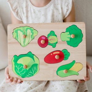 May include: Wooden vegetable puzzle with a lettuce, beetroot, broccoli, cabbage, carrot, and corn design. Each vegetable piece is brightly coloured and has a wooden peg for easy grasping. The puzzle is held by a person.