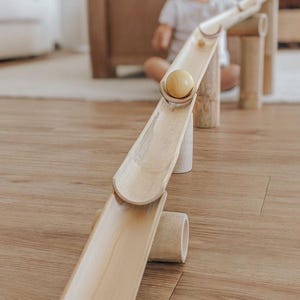 Può includere: Un gioco di biglie in legno con una finitura naturale. La pista è composta da più pezzi di bambù collegati e presenta una biglia che rotola lungo la pista.