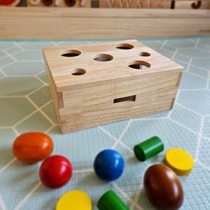 May include: Wooden toy box with a lid and six holes on top. The box is made of light wood and has a natural finish. There are seven colorful wooden shapes next to the box: a red ball, a blue ball, a yellow ball, a brown egg, a green cylinder, a yellow cylinder, and an orange oval.