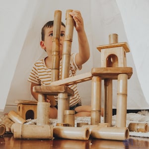 Può includere: Un bambino gioca con blocchi di legno naturale, costruendo una torre e un ponte. I blocchi sono cilindrici e rettangolari, di colore marrone chiaro. La scena è ambientata in interni, con uno sfondo bianco.