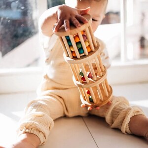 May include: A wooden toy with colorful beads that roll inside a cage. The toy is made of natural wood and has a simple design. It is a great toy for babies and toddlers to explore and learn.