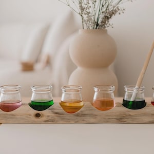 May include: A wooden paint palette with six small glass jars filled with colorful paints. A paintbrush rests in one of the jars. A beige vase with dried flowers sits behind the palette.