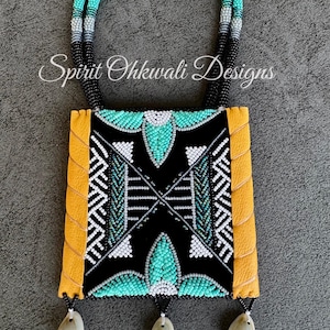 May include: A handcrafted beaded necklace with a black, turquoise, and white geometric design. The necklace features a square pendant with yellow leather edging and three shell pendants. The text "Spirit Ohkwali Designs" is visible.