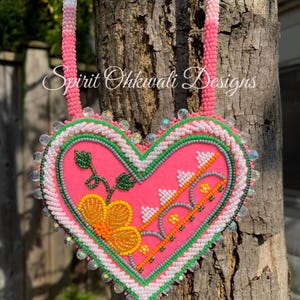 Pode incluir: Um coração de contas rosa com uma borda verde e branca, uma flor amarela com detalhes laranja e verde, e o texto "Spirit Ohkwali Designs".