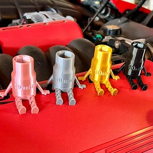 Puede incluir: Cuatro llaves de vaso de 10 mm en diferentes colores: rosa, gris, dorado y negro. Cada llave tiene forma de figura de dibujos animados con piernas y brazos. Las llaves están sobre una superficie roja.