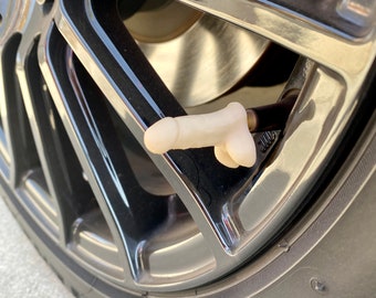 May include: A white plastic tire valve stem cap shaped like a human body. The cap is attached to a black tire valve stem on a black car tire.