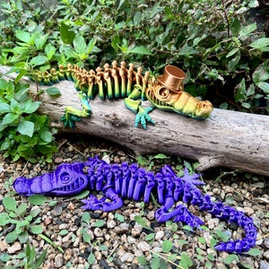 May include: Two articulated skeleton crocodile figurines, one is green and gold with a top hat, the other is purple, both are posed on a wood log and gravel.