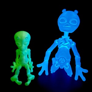 Puede incluir: Dos figuritas de extraterrestres que brillan en la oscuridad. El extraterrestre más grande es azul y tiene un diseño esquelético detallado. El extraterrestre más pequeño es verde y tiene un diseño más simple.