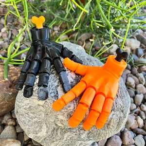 Puede incluir: Dos manos articuladas de plástico, una negra y otra naranja, con puntas de los dedos en forma de hueso. Las manos están posadas como si se dieran la mano.