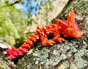 Può includere: Una statuetta di drago stampata in 3D di colore rosso e arancione con un corpo testurizzato e spinoso. Il drago è posato su un ramo d'albero ricoperto di muschio verde.