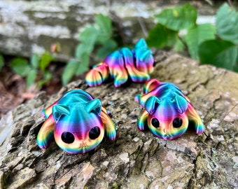 May include: Three colorful, rainbow-hued cat figurines with large black eyes are displayed on a textured, brown surface. The cat figurines are in various poses, with one standing and two lying down. The vibrant colors include shades of blue, green, yellow, and purple.