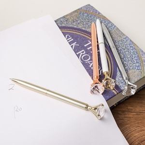May include: Four pens with clear diamond-shaped tops. The pens are gold, rose gold, silver, and white. The pens are lying on a book with the text "The Silk Road" visible.