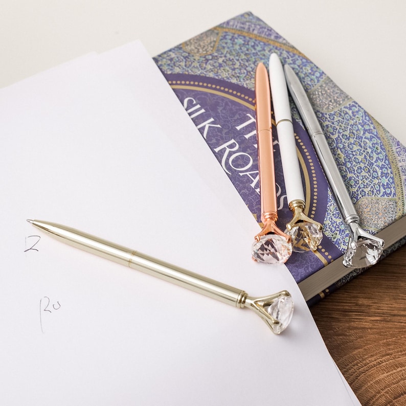 May include: Four pens with clear diamond-shaped tops. The pens are gold, rose gold, silver, and white. The pens are lying on a book with the text "The Silk Road" visible.