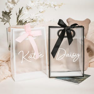 Könnte beinhalten: Zwei transparente Geschenktüten aus Plastik mit weißen Henkeln und weißen Schriftzügen. Die Tüte links hat ein rosa Band und steht "Kate". Die Tüte rechts hat ein schwarzes Band und steht "Daisy".