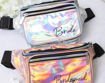 May include: Two iridescent fanny packs with black straps. The silver fanny pack says "Bride" in black script. The pink fanny pack says "Bridesmaid" in black script.