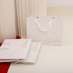 May include: Two white paper shopping bags with gold lettering. The bag on the left says "Kachira" and the bag on the right says "Kennetora".