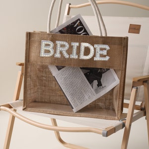 Könnte beinhalten: Eine natürliche Tragetasche aus Jute mit dem Wort "BRIDE" in glitzernden, silberfarbenen Buchstaben. Die Tasche hat eine transparente Netztasche und Seilgriffe. Zeitschriften sind im Inneren zu sehen. Die Tasche steht auf einem Klappstuhl.