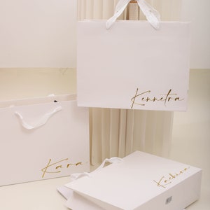 May include: Three white paper shopping bags with gold lettering. The bags are labelled "Kara", "Kennetra", and "Kachira".