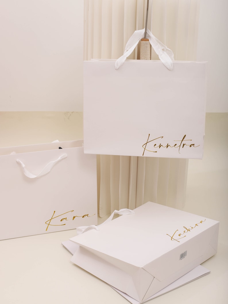 May include: Three white paper shopping bags with gold lettering. The bags are labelled "Kara", "Kennetra", and "Kachira".