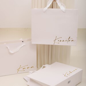 May include: Three white paper shopping bags with gold lettering. The bags are labelled "Kara", "Kennetra", and "Kachira".