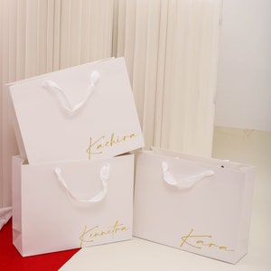 May include: Three white paper shopping bags with gold lettering. The bags are labelled "Kachira", "Kennetka", and "Kara".