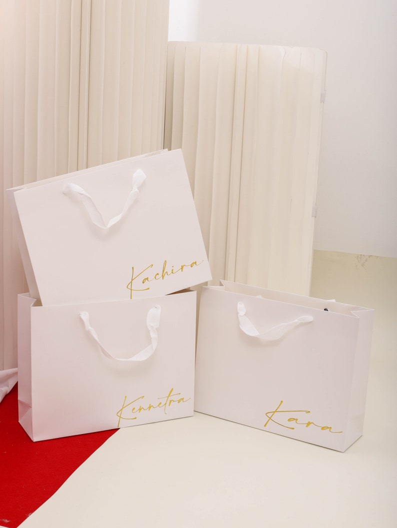 May include: Three white paper shopping bags with gold lettering. The bags are labelled "Kachira", "Kennetka", and "Kara".