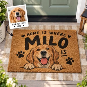 May include: A rectangular tan coir doormat featuring a printed illustration of a golden retriever with its paws resting on the bottom edge. The text reads "HOME IS WHERE MILO IS" in black lettering, accented by two black hearts and two paw prints.